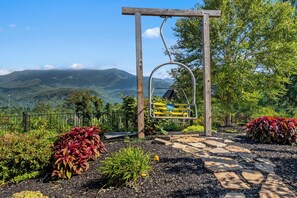 Um baloiço de madeira com estrutura de metal, rodeado por canteiros ajardinados com diversas plantas, um caminho de pedra e uma paisagem montanhosa ao fundo.