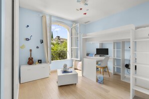 A room with a bed, a desk with a computer, a window with curtains, and a view of buildings outside. - Villa Jasmin | French Riviera Retreat (Vence)