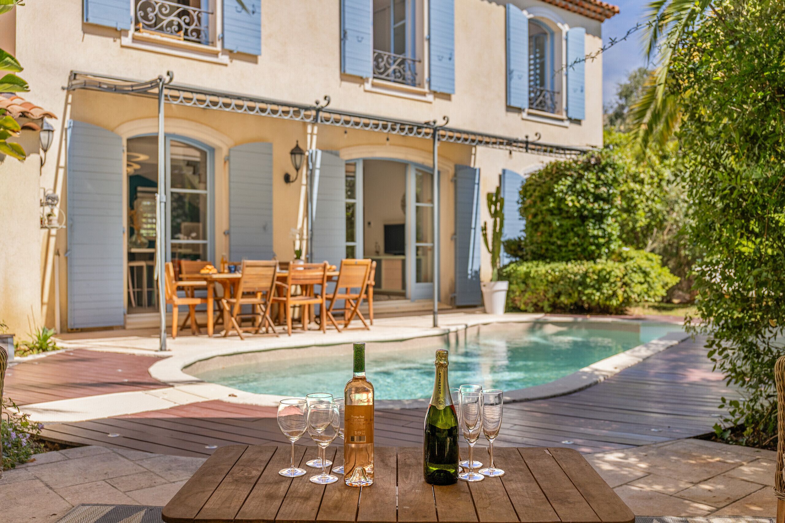 A poolside setting with a wooden table, a bottle of champagne, and glasses.