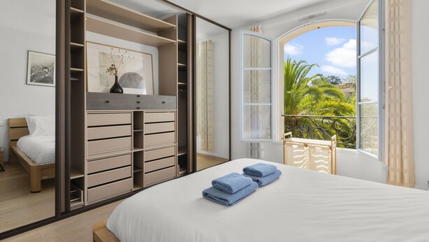 Ein Schlafzimmer mit Bett, Kleiderschrank, einem Fenster mit Blick ins Grüne und einem Balkon mit Sitzgelegenheit.