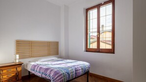 Un dormitori amb un llit de fusta, una petita taula de nit, una finestra amb persianes i vista a edificis a l'exterior.