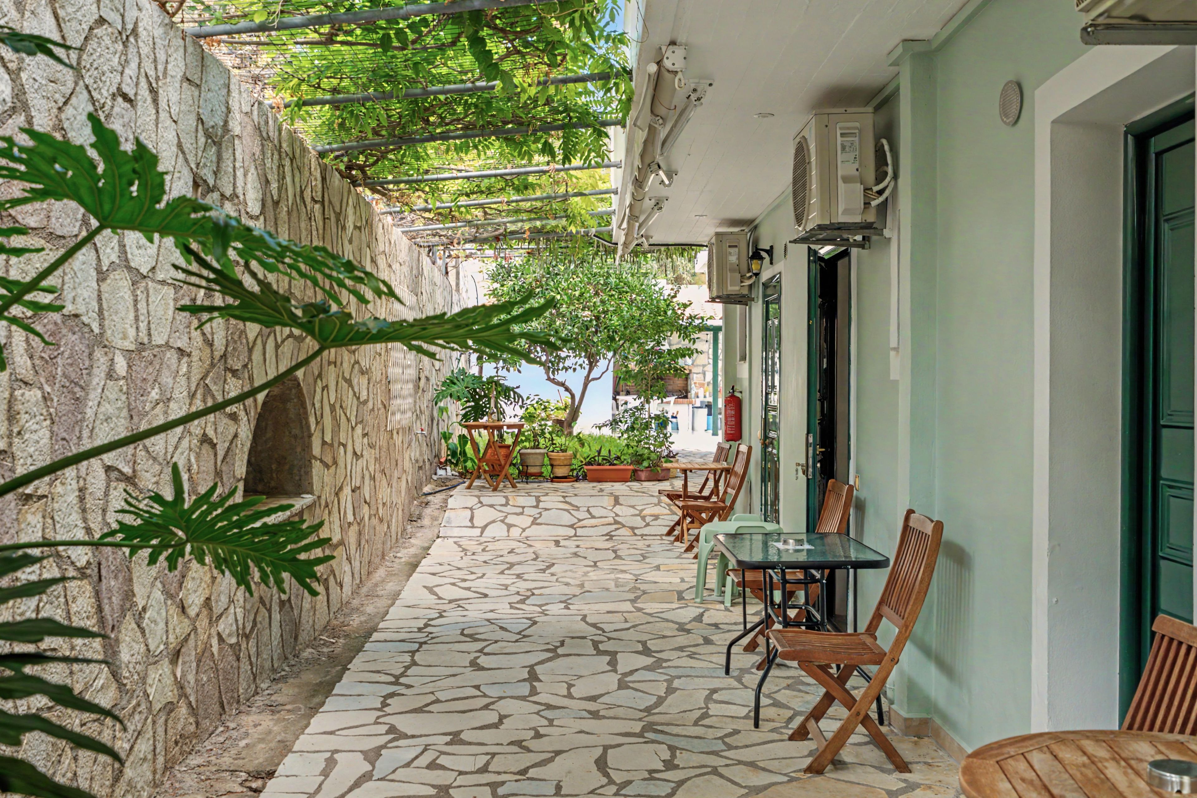 Un espacio al aire libre con suelo de piedra, mesas y sillas, una pérgola con plantas trepadoras y un edificio con puerta verde.