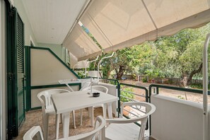 Un espace extérieur aménagé en terrasse, avec une table et des chaises blanches, un coin salon abrité et une vue sur des arbres et une clôture.