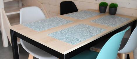 A dining table with four patterned placemats, two chairs with teal upholstery, and two potted plants on the wall.