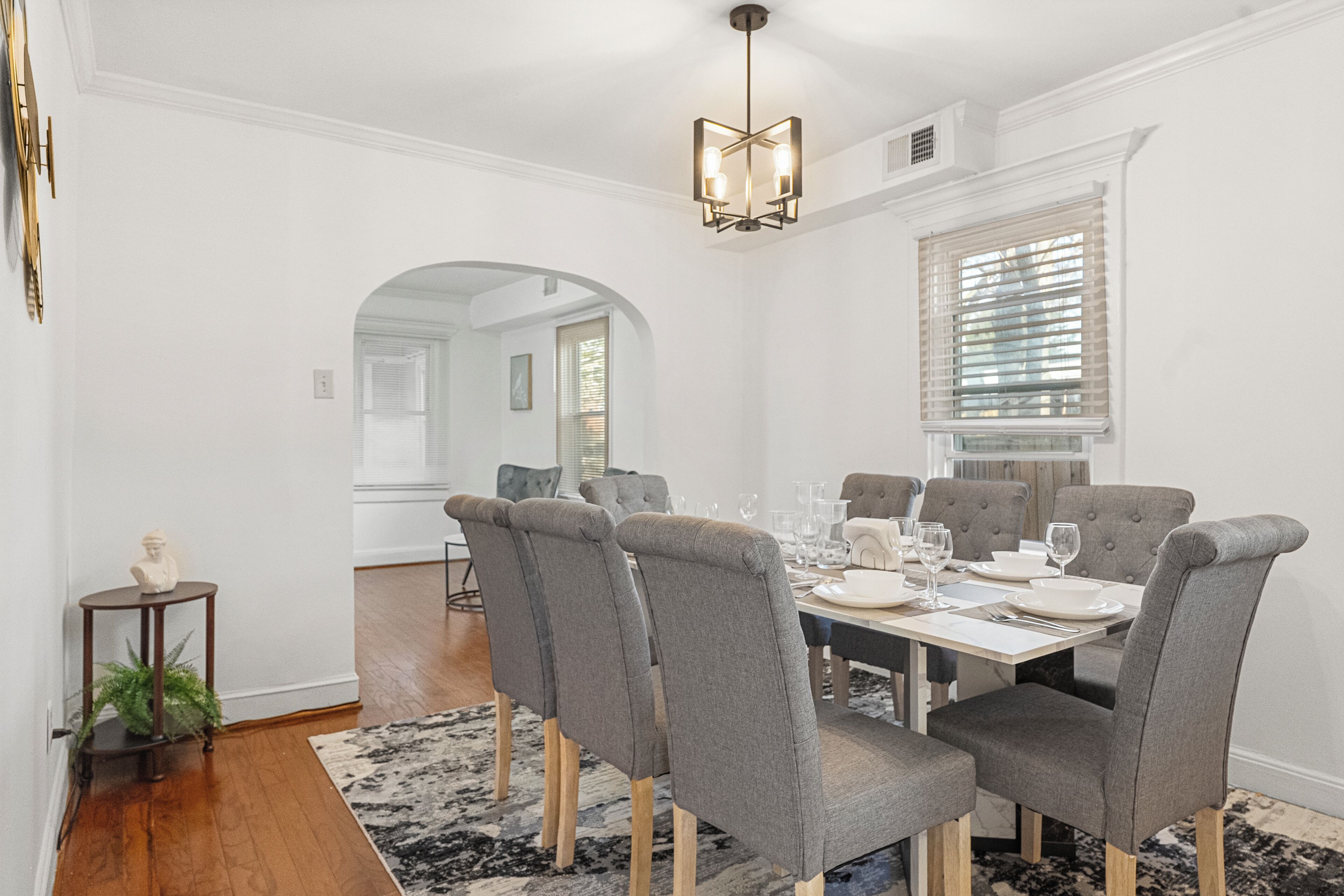Ein Esszimmer mit einem rechteckigen Tisch, gedeckt für sechs Personen, umgeben von sechs grauen Polsterstühlen mit Holzbeinen. Eine moderne Chandeliebre hängt von der Decke, und ein großes Fenster mit Jalousien ist an der rechten Wand zu sehen.