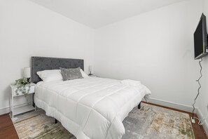 Ein Schlafzimmer mit einem weißen Bett, grauem Kopfteil, Nachttischen mit Lampen und einem an der Wand montierten Fernseher.