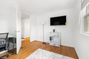 Ein modernes, gut beleuchtetes Zimmer mit Holzboden, einem Schreibtisch mit Stuhl, einer Stehlampe, einem an der Wand montierten Fernseher und einem Bücherregal.