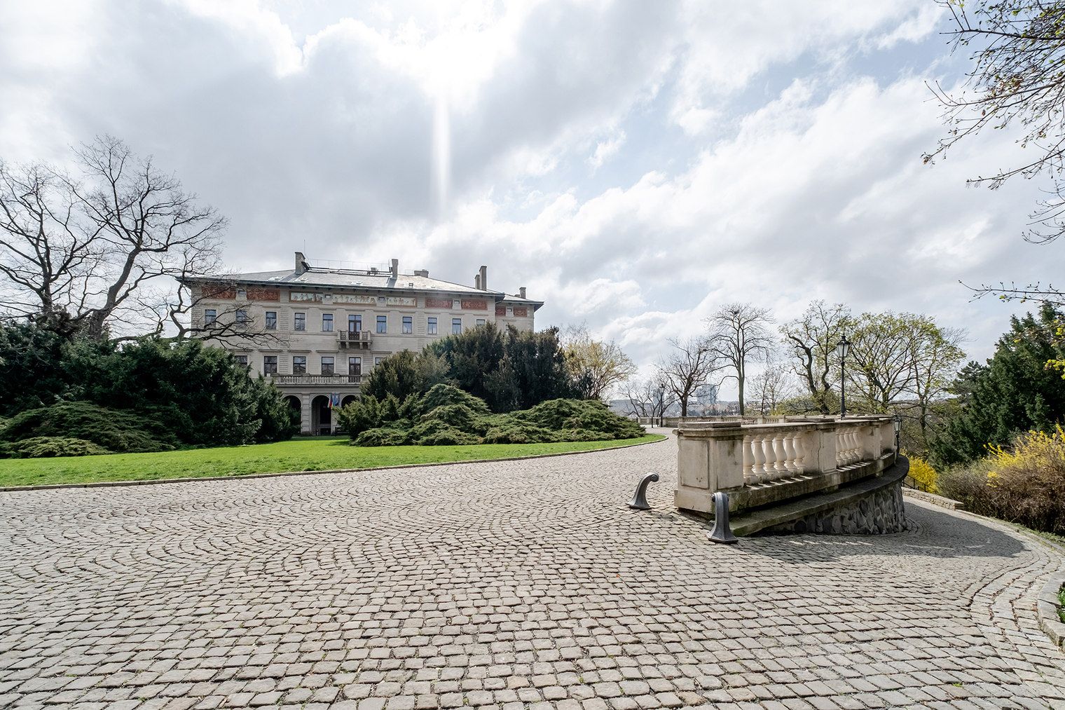 Une cour pavée de pierres, avec une fontaine, un grand bâtiment en arrière-plan et une pelouse bien entretenue.