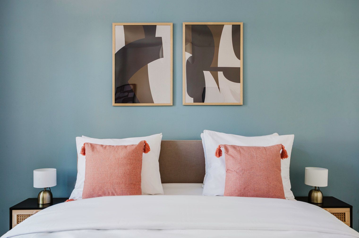 A neatly made bed with white and pink pillows, two abstract paintings on the wall, and bedside lamps on either side.