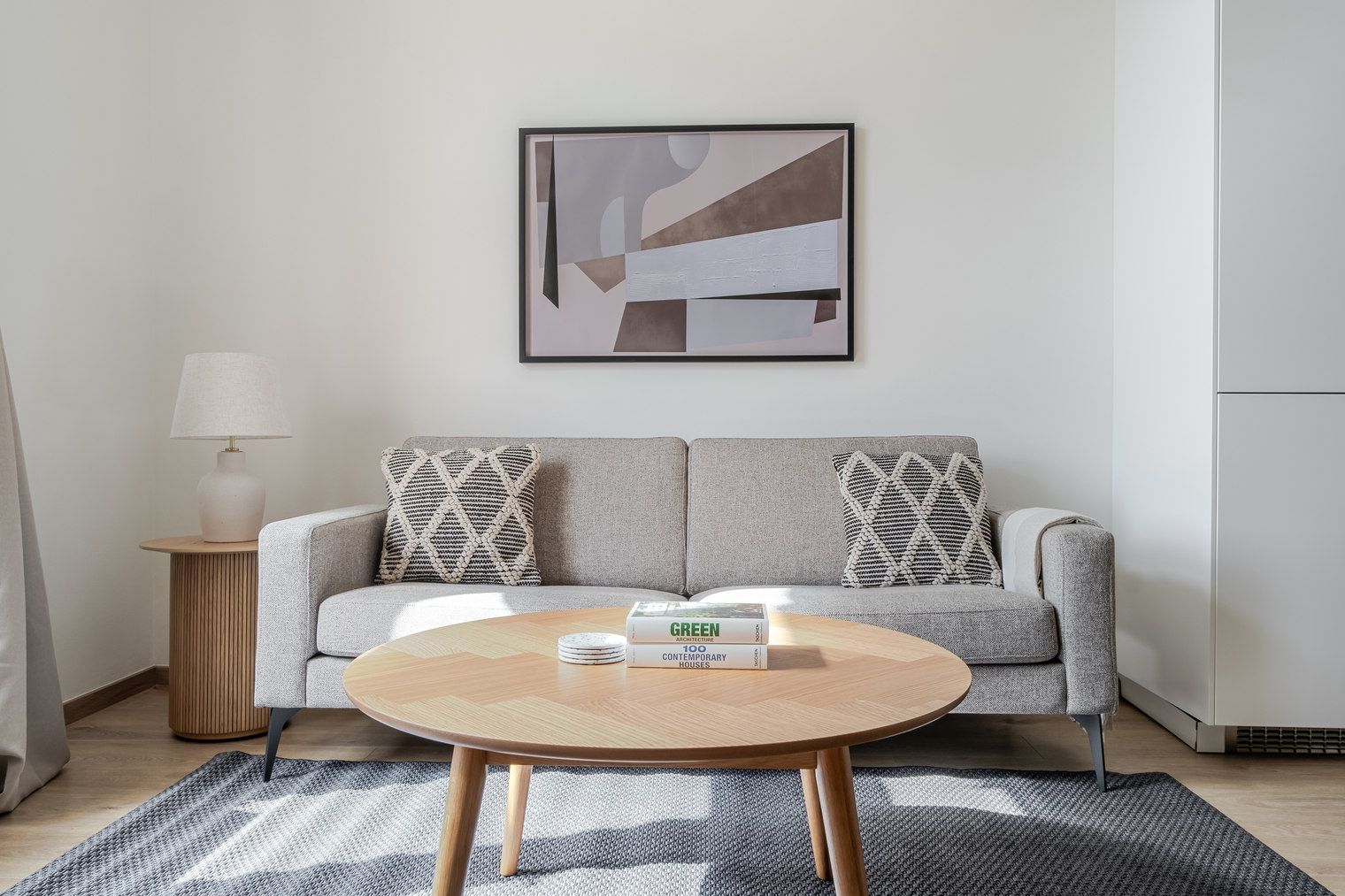 A modern living room with a grey sofa, two patterned throw pillows, a round wooden coffee table, and a framed abstract painting on the wall.