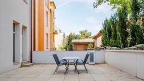 Um pátio externo com mesa e cadeiras, uma parede branca e um edifício com fachadas laranja e branco ao fundo.