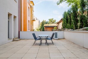 Eine Terrasse im Freien mit Tisch und Stühlen, einer weißen Wand und einem Gebäude mit orange-weißen Fassaden im Hintergrund.
