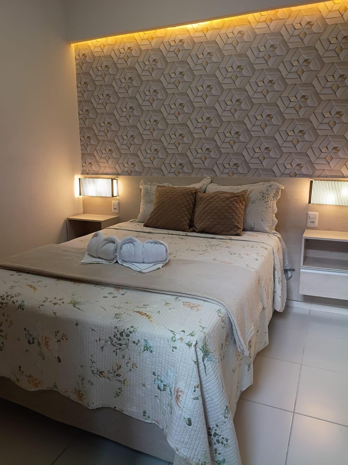 Habitació d'hotel amb un llit gran, un panell de capçal amb dibuixos, làmpades laterals i un edredó floral.