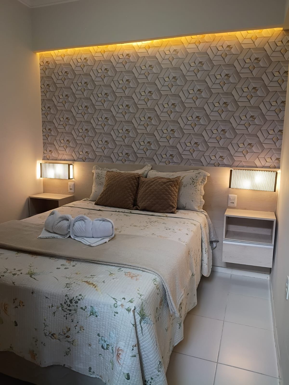 Una habitació d'hotel amb un panell de capçalera estampat, un llit amb roba de llit floral, dos almoines i làmpades laterals.