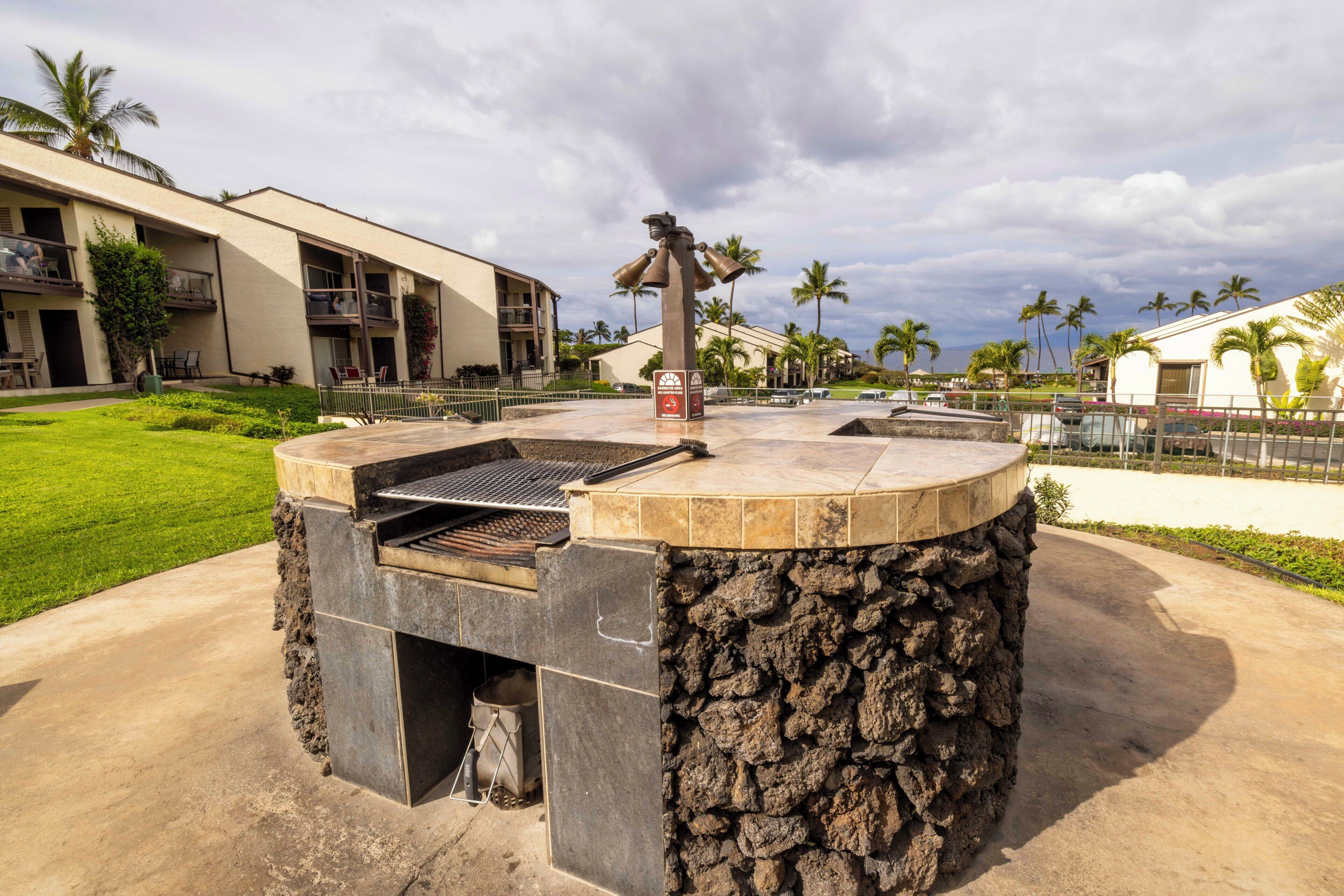 An outdoor barbecue area with a stone structure, surrounded by a fenced property with a pool and palm trees.