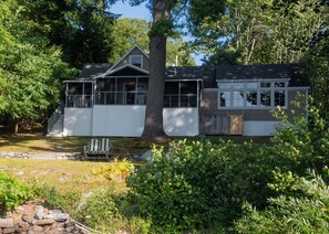 A two-story house with a covered porch, a visible garage, and a stone retaining wall. - Historic 3-BR cottage on coveted Megunticook Lake with AC/heat in Camden ME. (Camden)