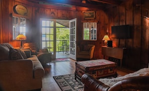 A cozy room with a wooden interior, a sofa, a small table, a television, and a window with a view of greenery. - Historic 3-BR cottage on coveted Megunticook Lake with AC/heat in Camden ME. (Camden)