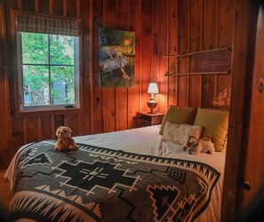 Ein Zimmer mit Holzwänden, einem Bett mit gemustertem Überwurf, einem Fenster mit Blick auf Grünflächen, einem Gemälde und einer Lampe.