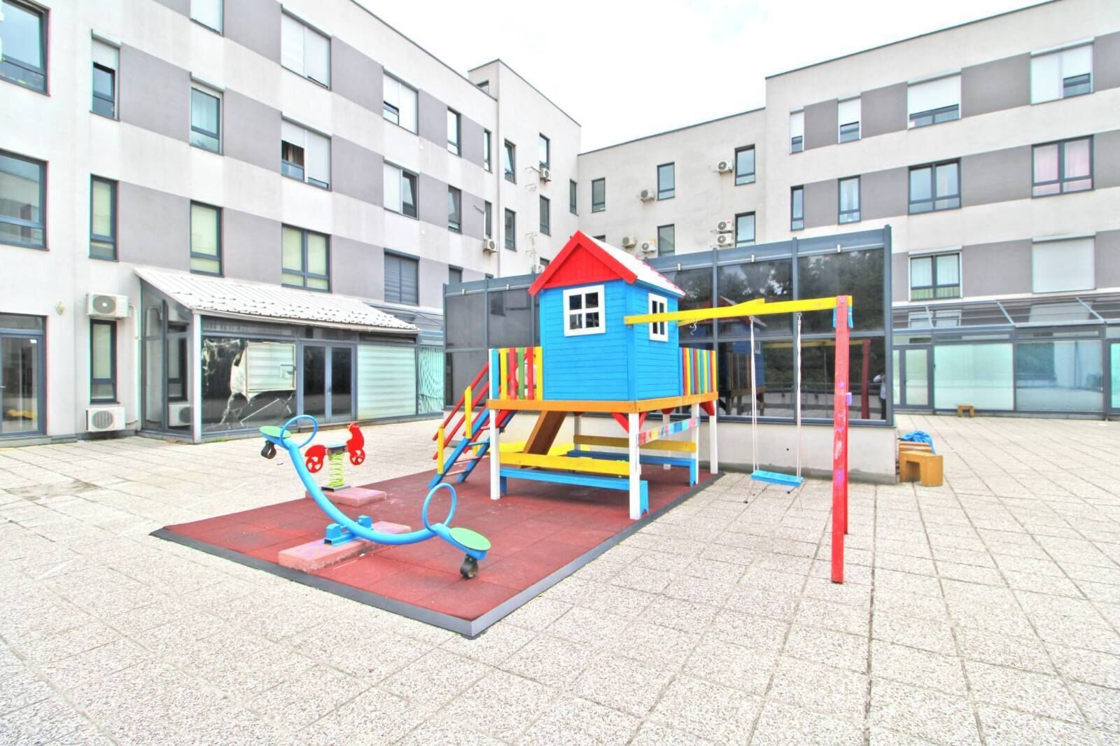 Une aire de jeux avec un toboggan et des balançoires, entourée d’un bâtiment doté de grandes fenêtres et de climatiseurs.