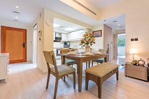A modern kitchen with white cabinets, a dining area with a wooden table and chairs, and a small side table with a lamp. - Quiet 2BR Haven Steps from The Greens Lakes and Emirates Golf Club,Al Dhafrah 2 (Dubai)