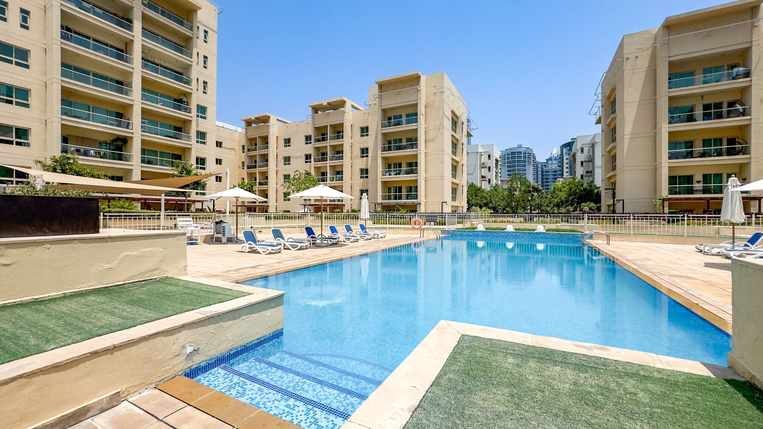 Una piscina con sillones de descanso y sombrillas, rodeada de edificios de varios pisos.