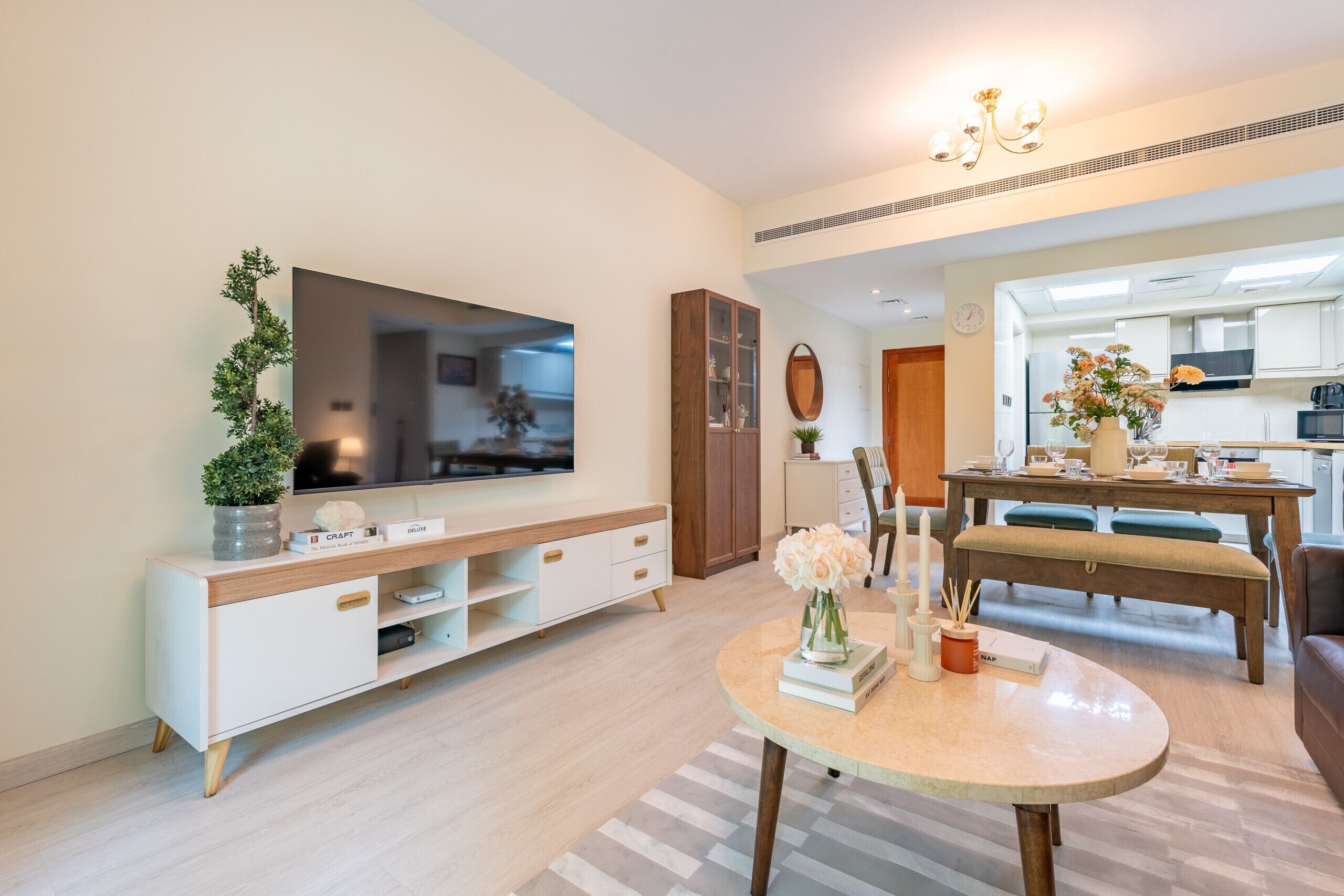 A modern living room with a flat-screen TV, a wooden cabinet, a dining table with chairs, and a kitchen in the background.