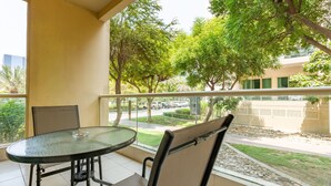 A balcony with a glass table and two chairs, overlooking a street with trees and parked cars. - Quiet 2BR Haven Steps from The Greens Lakes and Emirates Golf Club,Al Dhafrah 2 (Dubai)