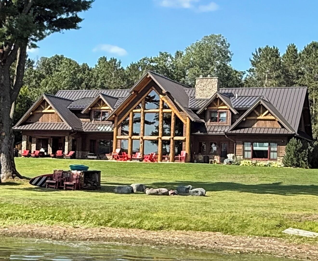 Une grande cabane en rondins, dotée d’une tour centrale imposante, d’un porche couvert avec des chaises rouges et d’une cour verdoyante où se trouvent un bateau et des rochers.