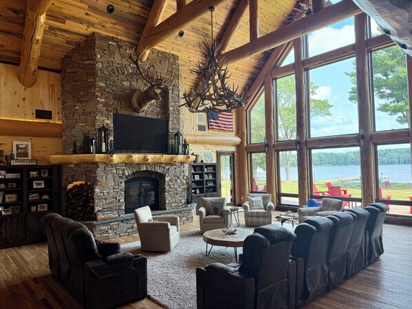 A spacious living room with a stone fireplace, leather sofas, and a large window overlooking a lake.