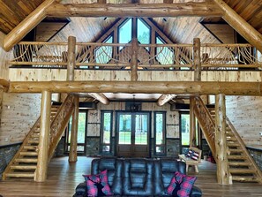 A spacious interior with a high ceiling, wooden beams, a fireplace, a leather sofa, and a staircase. - Spacious 5-bedroom lodge on Bone Lake with 2 guest cabins, pickleball and sauna. (Milltown)