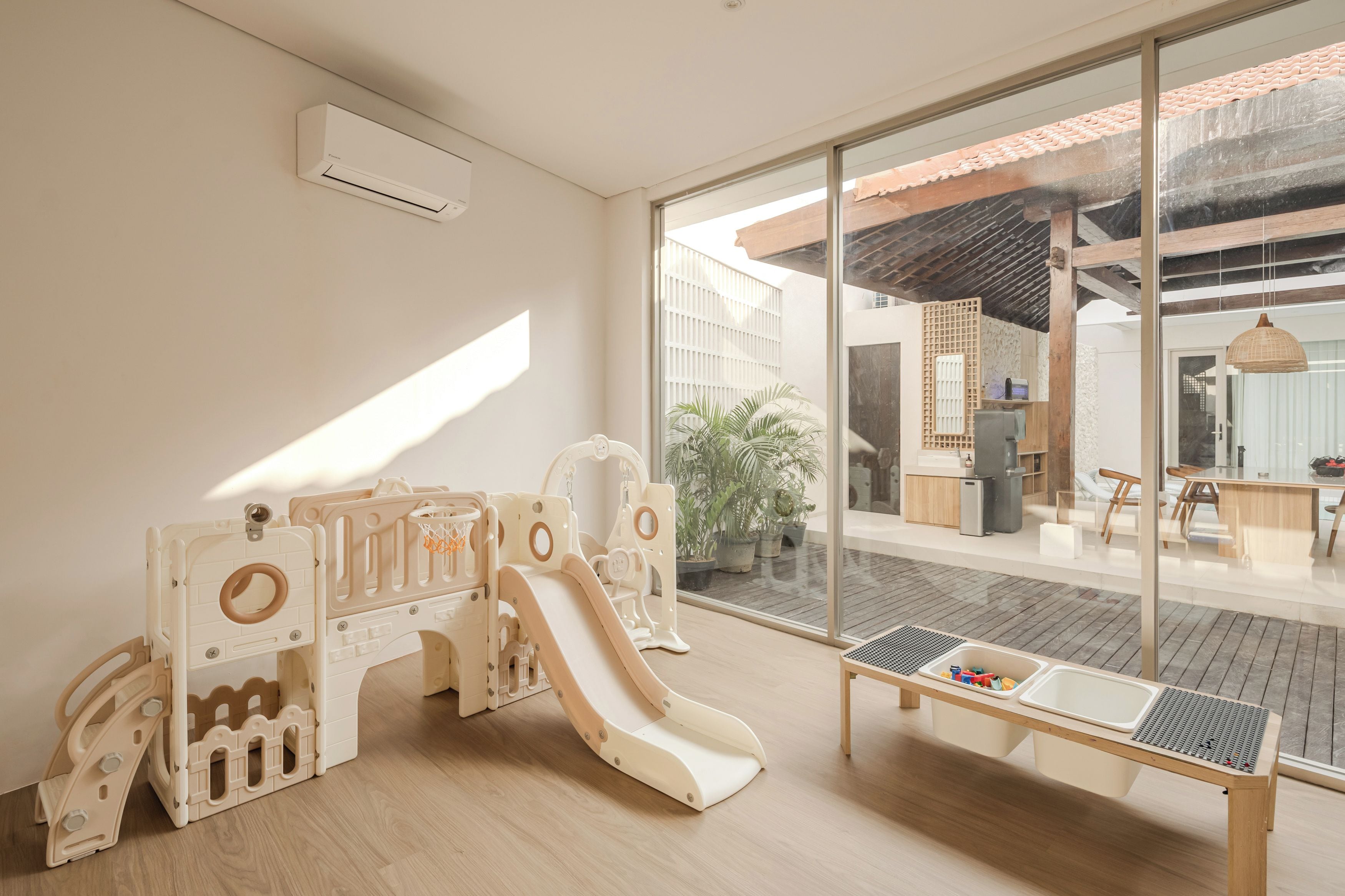 Un espace de jeux pour enfants avec un toboggan et des structures ludiques, une petite table avec des jouets, et une vue sur l’extérieur grâce à de grandes fenêtres.
