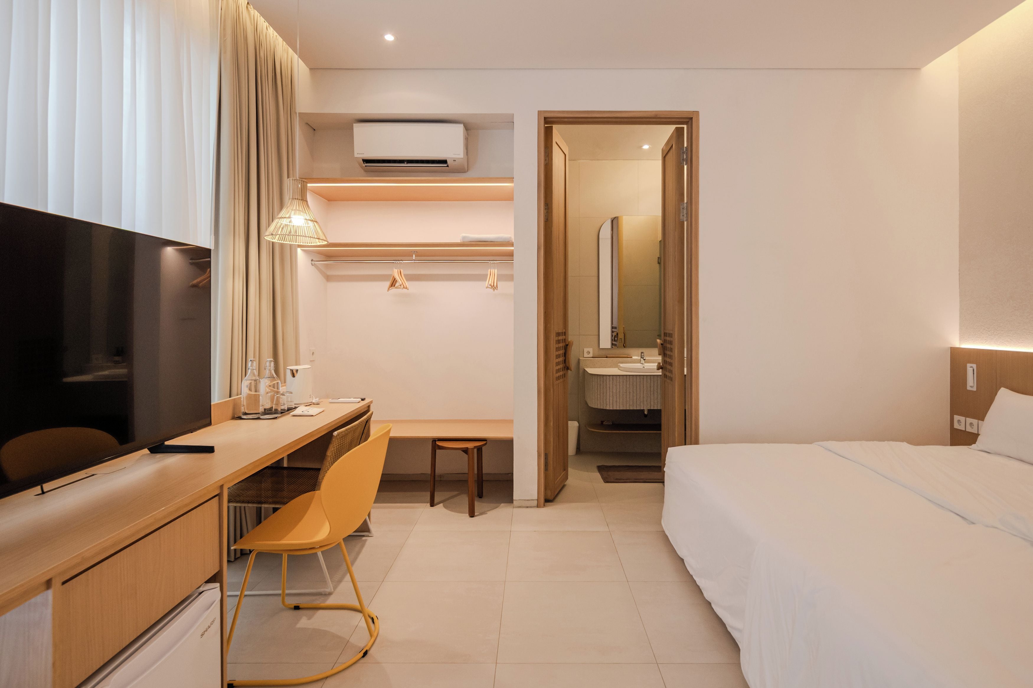A hotel room with a bed, a desk, a chair, a TV, a mirror, and a bathroom visible through an open door.