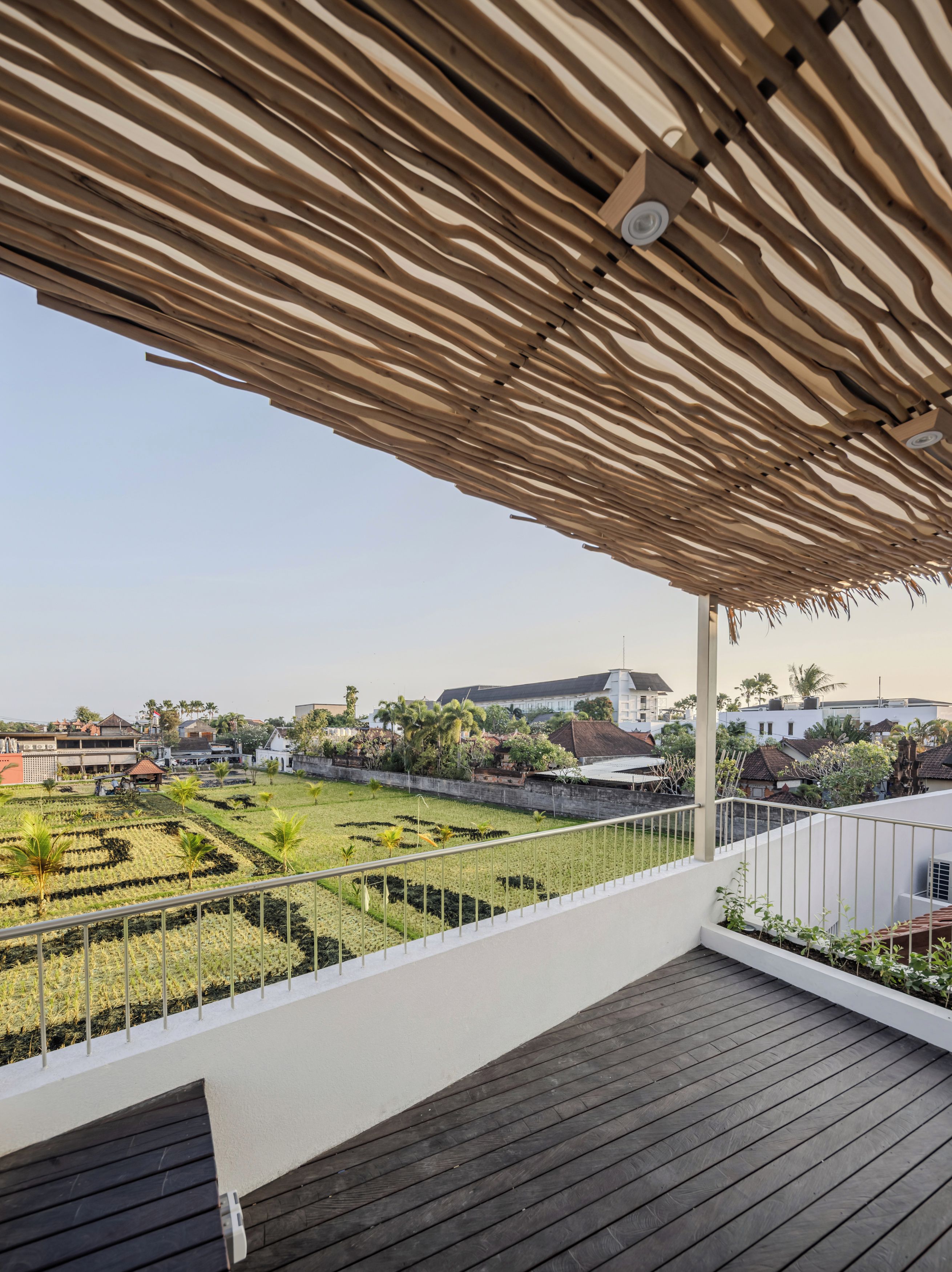 Eine Dachterrasse mit Holzdeck, einem Reetdach und Blick auf einen angelegten Garten mit Palmen.