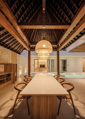 A spacious dining area with a long table and chairs, a hanging light fixture, and a stone wall.
