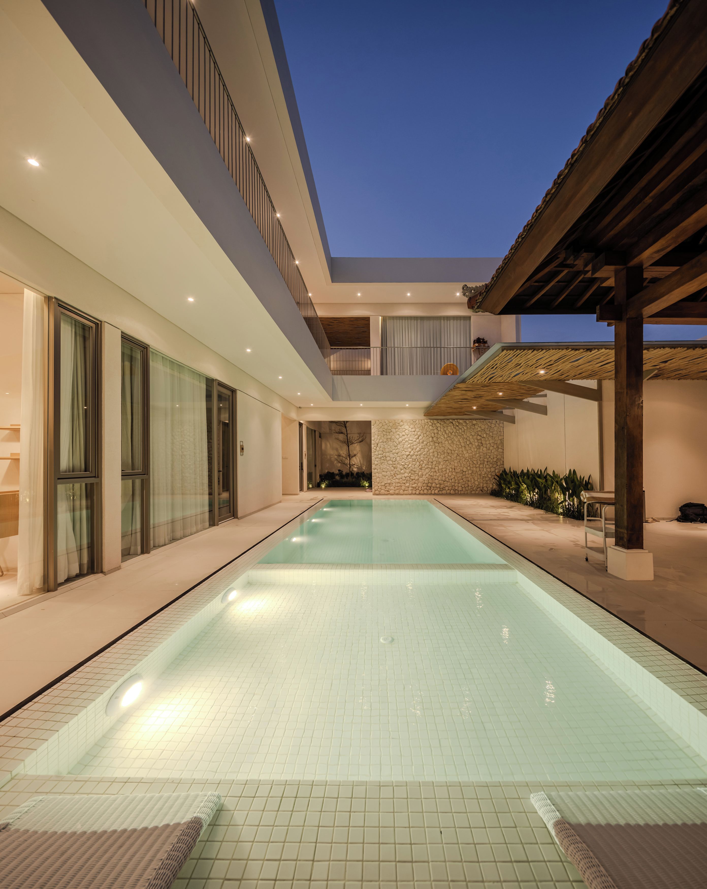 A modern indoor swimming pool with a clear view of the sky, surrounded by a spacious area with a wooden pergola and a stone wall.