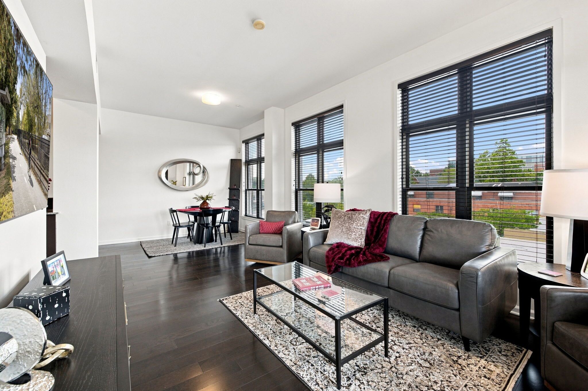 A modern living room with a dark wood floor, a glass coffee table, a grey sofa, and a dining area with a red tablecloth.