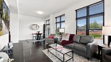 A modern living room with a dark wood floor, a glass coffee table, a grey sofa, and a dining area with a red tablecloth.