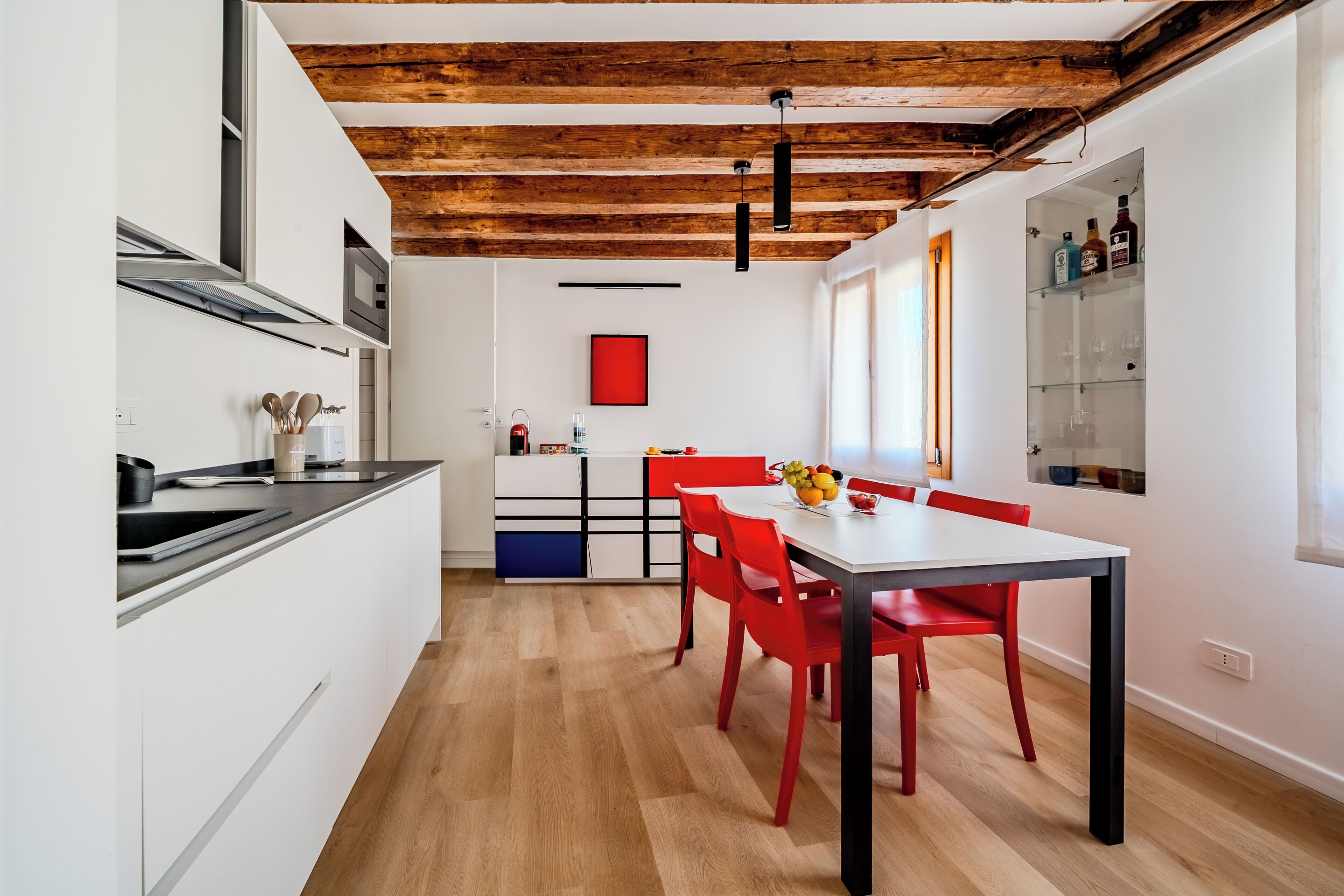 A modern kitchen with white cabinetry, a wooden ceiling, and a dining area with red chairs and a white table.