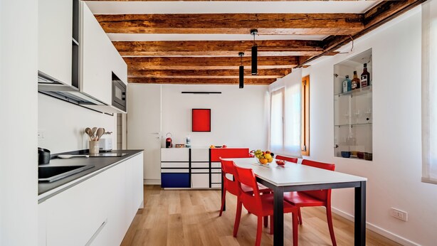 A modern kitchen with white cabinetry, a wooden ceiling, and a dining area with red chairs and a white table. - Suite Mondrian 100m from Biennale - Canal View (Venezia)