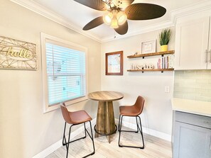 A kitchen with a round wooden table, two bar stools, a ceiling fan, and shelves with books and decorative items. - Cozy Cottages, Min 2 Beach, Shops & Restaurants! (San Diego)
