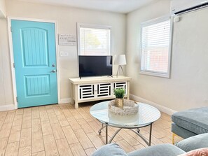 A room with a blue door, a flat-screen TV on a white console, a round glass table with a woven basket, and grey upholstered furniture. - Cozy Cottages, Min 2 Beach, Shops & Restaurants! (San Diego)