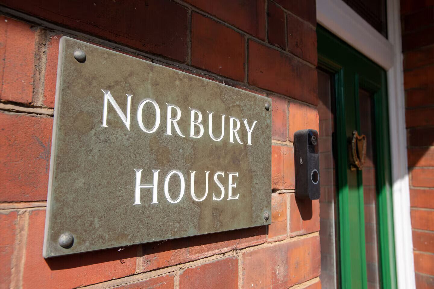 Une plaque en métal indiquant « NORBURY HOUSE », fixée à un mur de briques, avec une porte verte visible en arrière-plan.