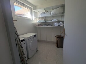 Una sala de lavandería con una lavadora, repisas con ropa doblada y un bote de basura.