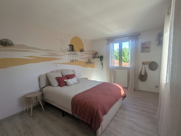 Un dormitorio con cama, una mesita redonda, una ventana con cortinas y un mural en la pared.