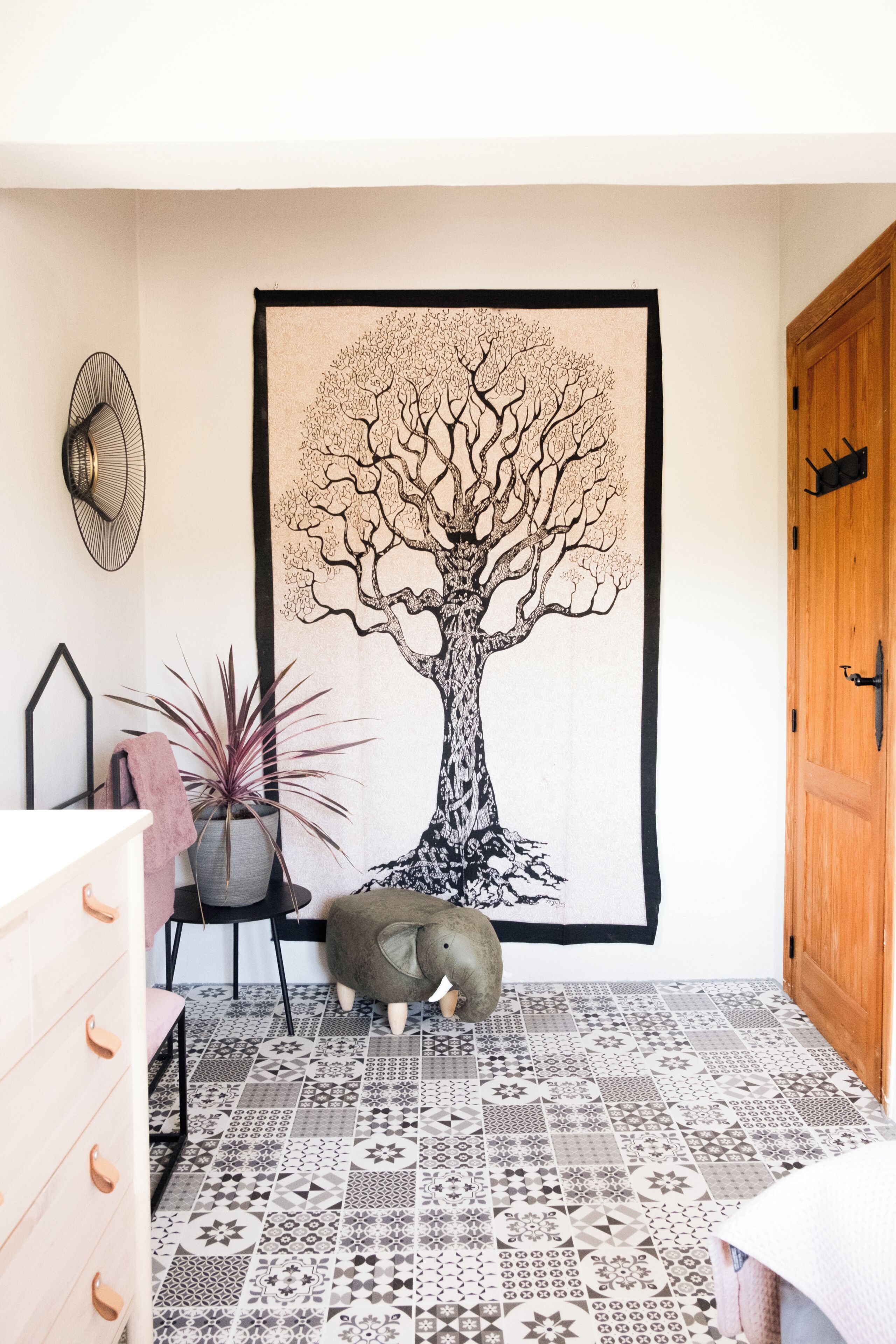 Een kamer met een groot wandtapijt van een boom, een potplant, een kleine olifantbeeldje en vloerbedekking met een patroon.