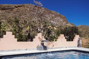 A swimming pool with a water feature, surrounded by a landscaped area with plants and a view of a mountainous landscape.