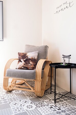 A wicker chair with a grey cushion and a cushion with a bird painting, a small black table with a statue, and a patterned floor.