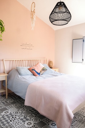 A bedroom with a bed, a bedside table, a hanging light fixture, and a wall with a dreamcatcher and a motivational quote.