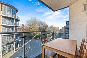 Un balcon avec une table et des chaises en bois, donnant sur une rue animée par la circulation des voitures et bordée d’immeubles.