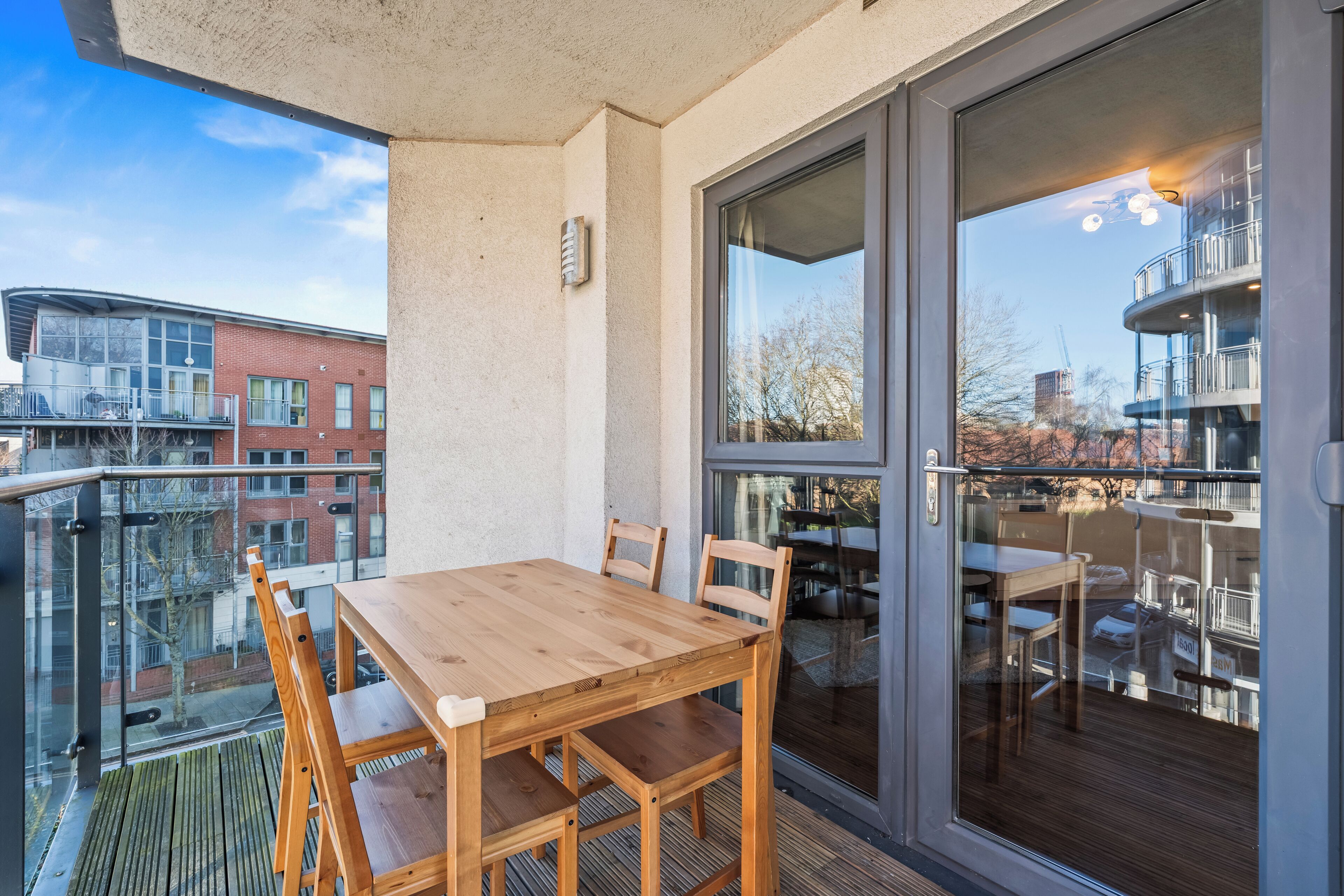 En balkon med et træbord og stole, en glasdør der fører til et udendørs område, og udsigt over en by med bygninger.